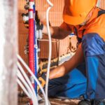 Caucasian Plumber Wearing Orange Hard Hat Installing Bathroom Water Supply Inside Newly Developed Concrete Blocks Building. Construction Site Theme.